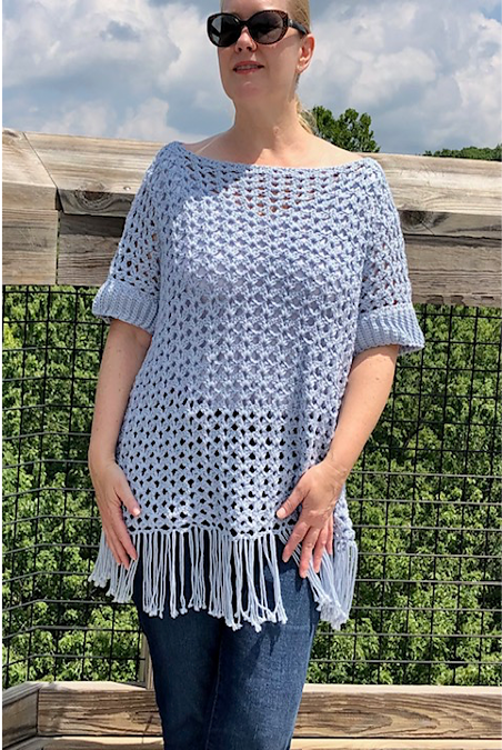 Woman wearing the Sea Shells Top, a light blue crochet poncho-style top with short sleeves and fringed hem